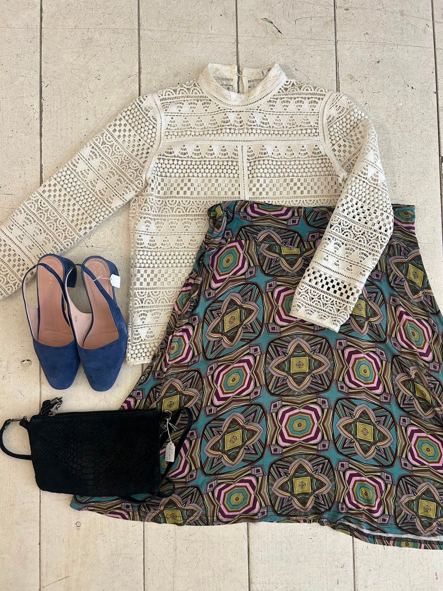 Flatlay met een crème kanten blouse, gedessineerde rok, blauwe suède muiltjes en een zwart schoudertasje.