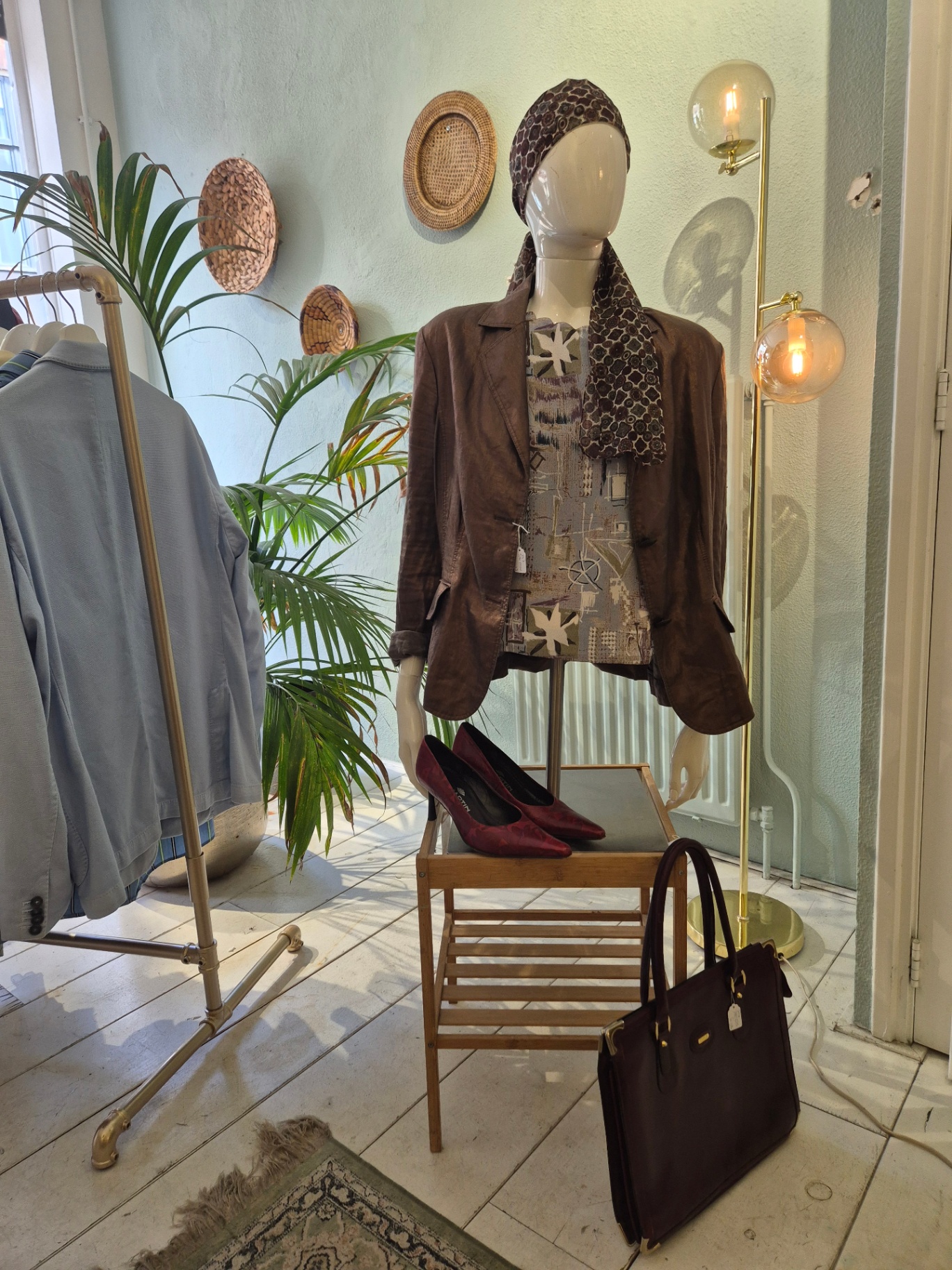 Etalagepop met bruin linnen blazer, sjaal om het hoofd en bordeauxrode pumps, naast een gouden staande lamp.