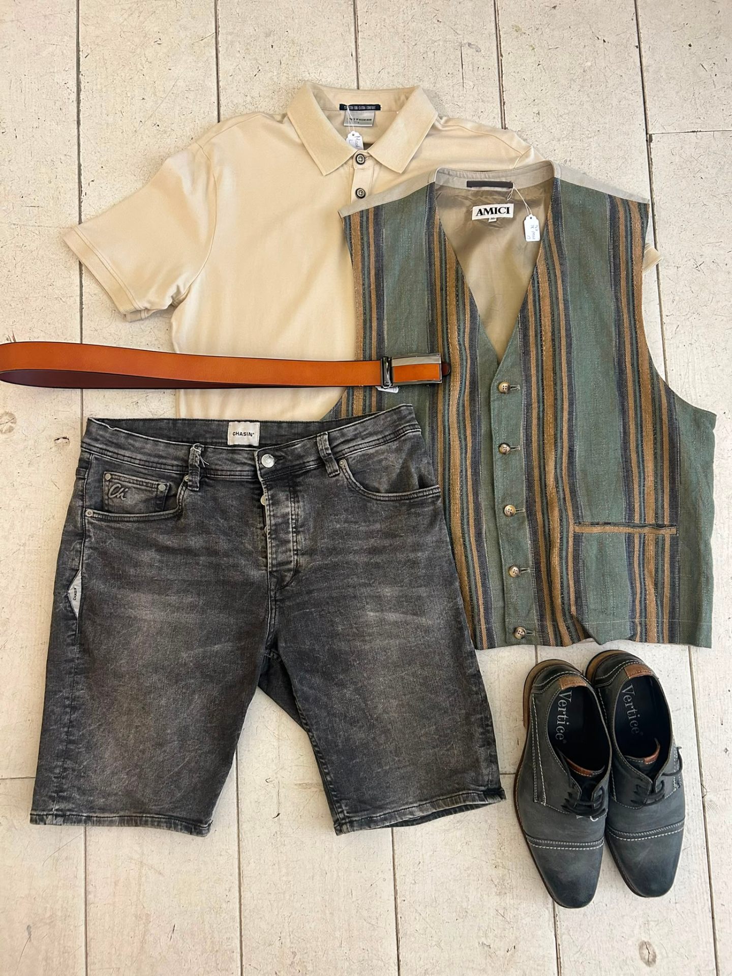 Herenflatlay met crème polo, gestreept gilet, grijze denim short, cognac riem en donkere veterschoenen.