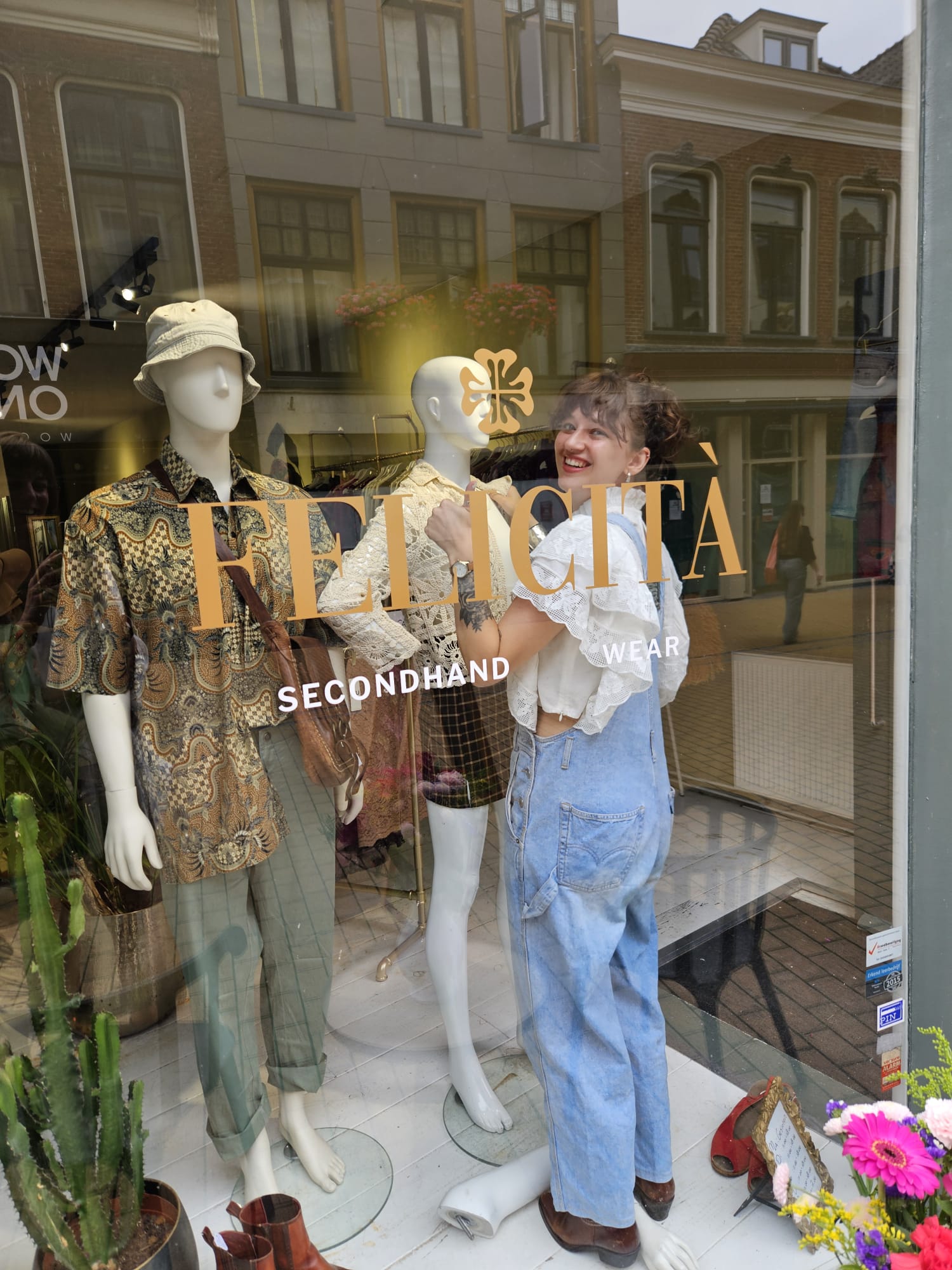 Medewerker van Felicità stijlt lachend een etalagepop, gezien vanaf de straat door de winkelruit met het gouden Felicità-logo.